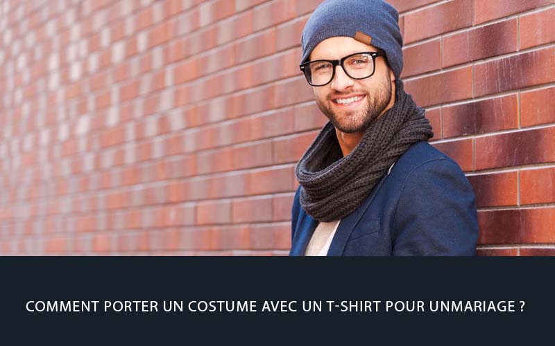 Homme souriant portant un costume bleu avec t-shirt blanc, bonnet et écharpe, look moderne et décontracté pour un mariage stylé et tendance