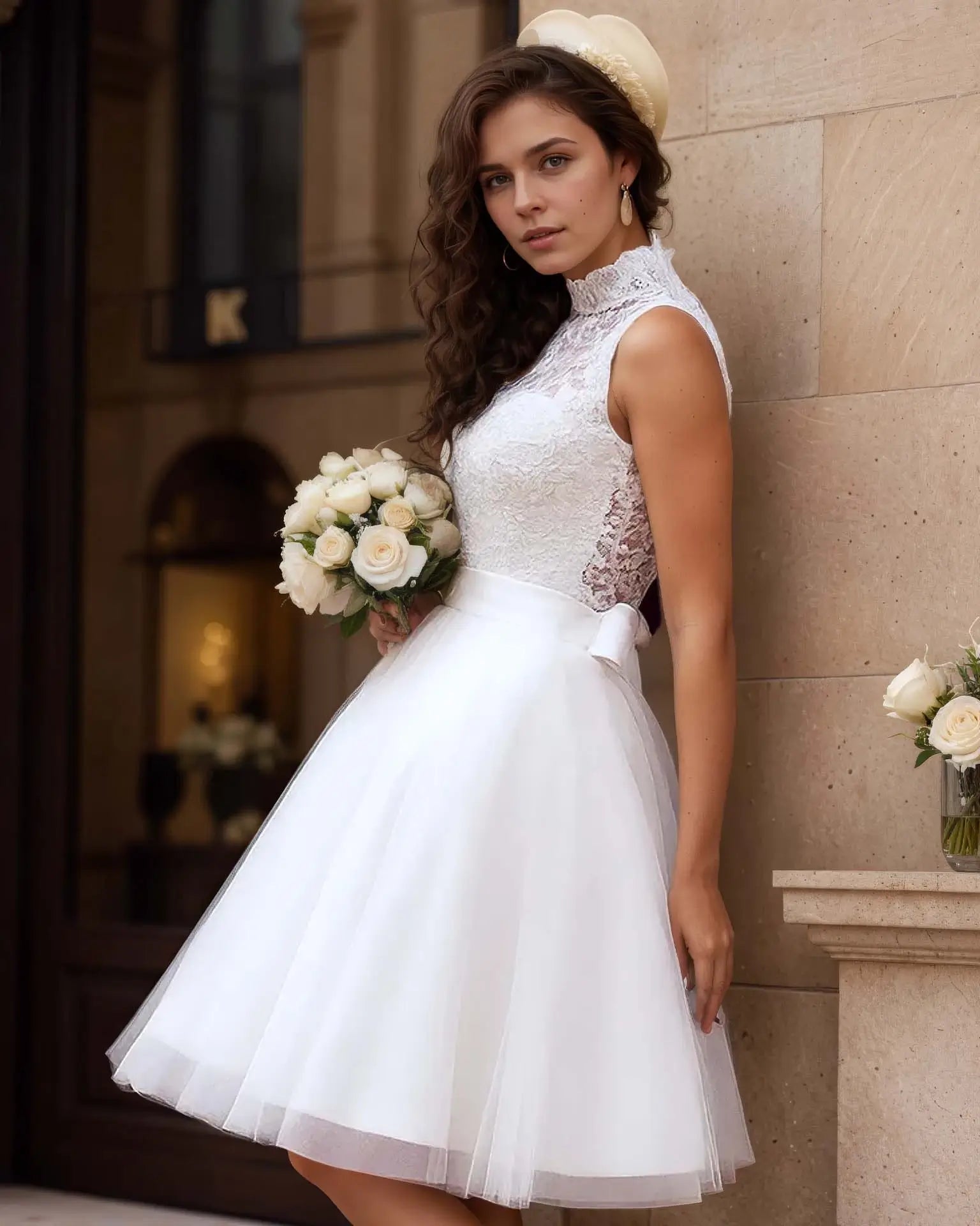 Robe de mariée courte vintage en dentelle avec jupe évasée, mariée tenant un bouquet blanc devant une façade élégante, style rétro chic.